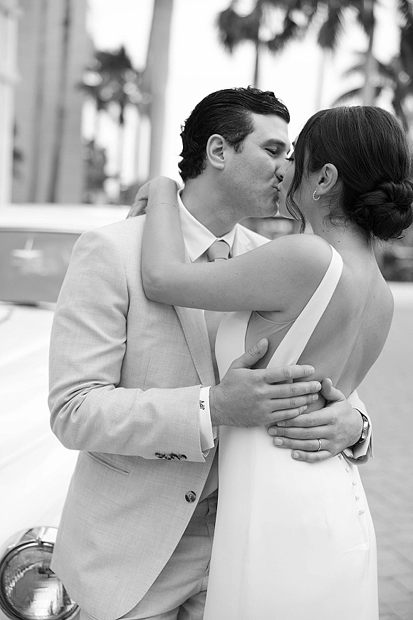 Wedding kiss portrait of bride and groom kissing close up beside a vintage car, palm trees and a street facade behind them