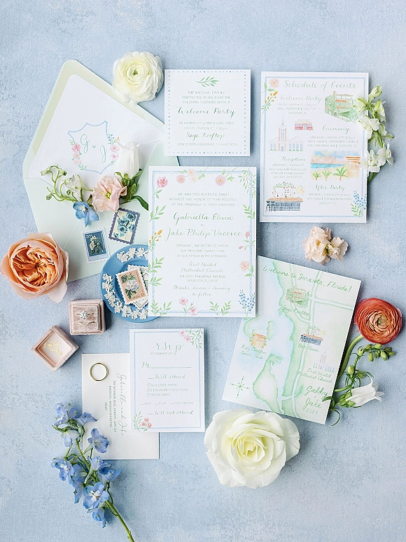 Invitation suite with watercolor wedding invitations, RSVP and map cards, wax-sealed envelopes, rings, and pastel flowers on light blue fabric backdrop