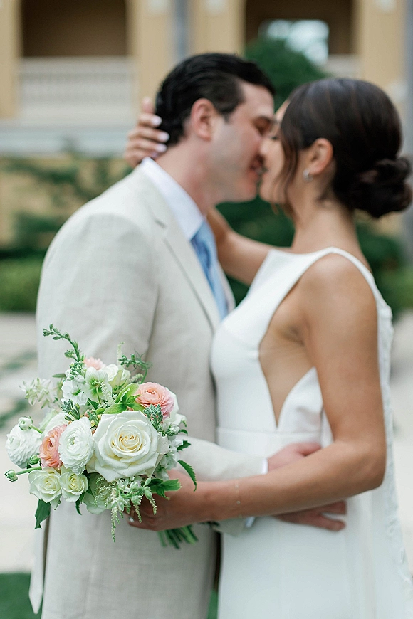 Wedding kiss as bride holds a white rose bouquet, embracing groom in a beige suit on stone steps in a garden with columns