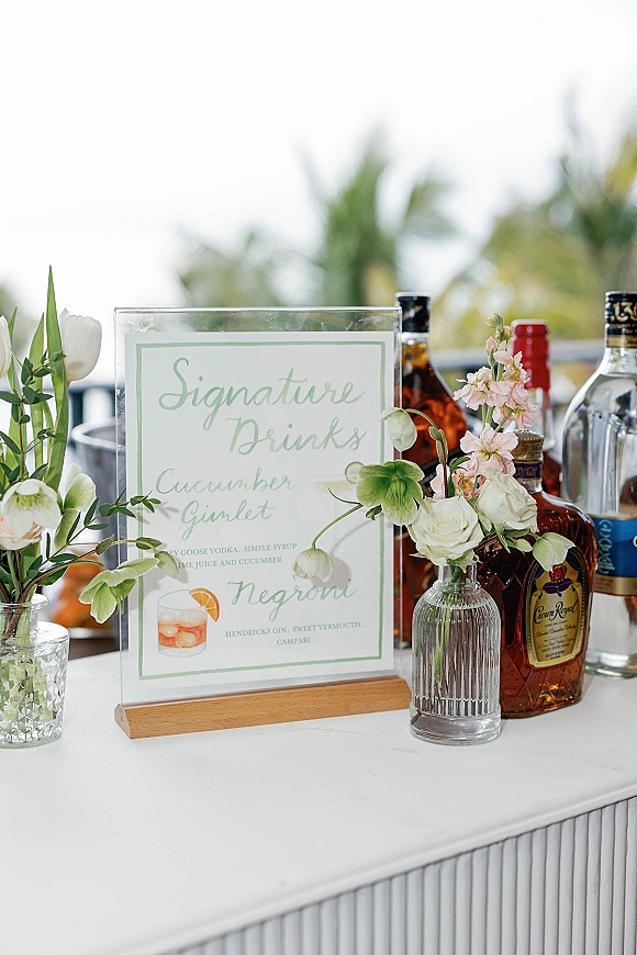 Signature drink sign wedding bar sign in an acrylic frame on a wooden base, with liquor bottles and bud vases of roses on a terrace