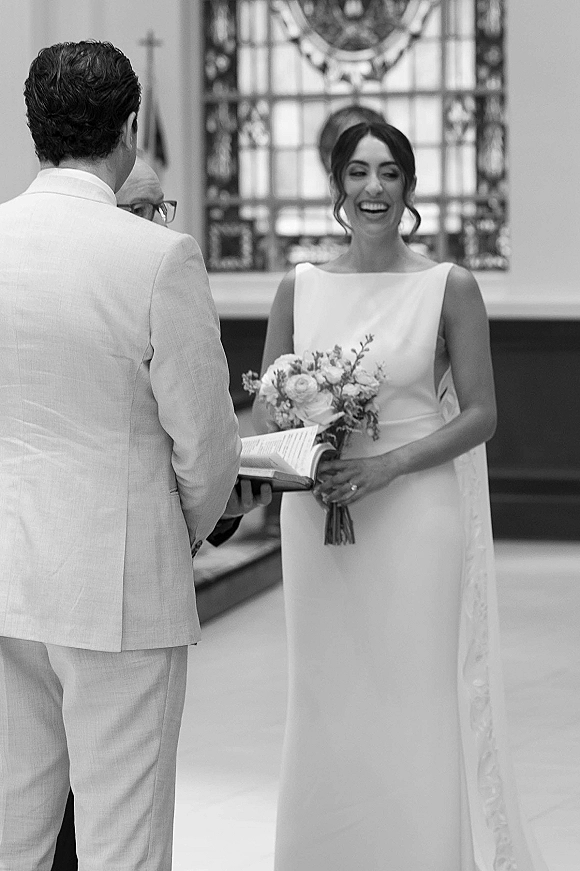 Wedding vows as bride laughs holding a simple bouquet with greenery beside groom in a light suit, inside a church with stained glass window