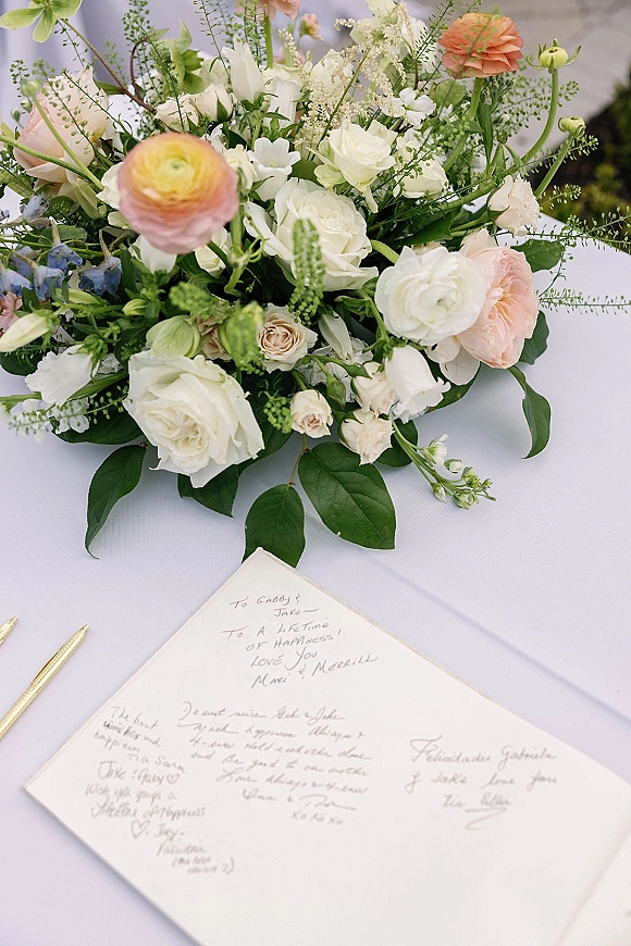Wedding guestbook on a wedding guestbook table with a gold pen beside a white rose centerpiece, open to handwritten messages on a white cloth