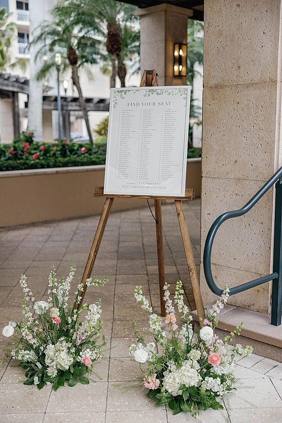 Wedding seating chart on a wooden easel with white and blush floral arrangements and greenery along a palm-lined outdoor walkway