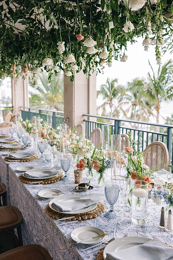Reception tablescape with an outdoor reception table set in blue toile linens, pastel centerpieces, taper candles, and hanging greenery on an ocean-view terrace