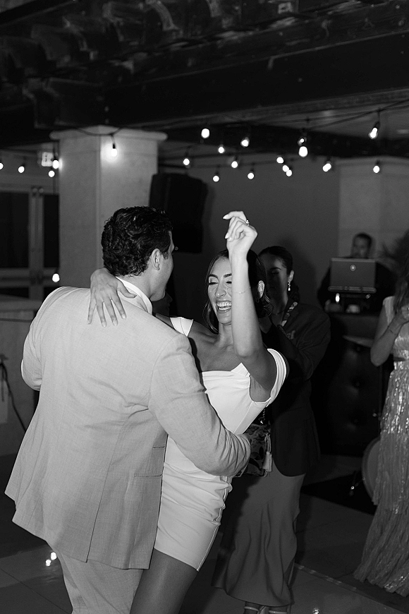 Wedding reception dancing as the bride and groom dance under string lights on an indoor floor, guests and DJ booth behind, bride in off-the-shoulder dress
