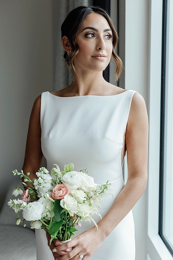 Bridal portrait of a bride holding bouquet of white roses and ranunculus in a simple white wedding dress by soft window light indoors