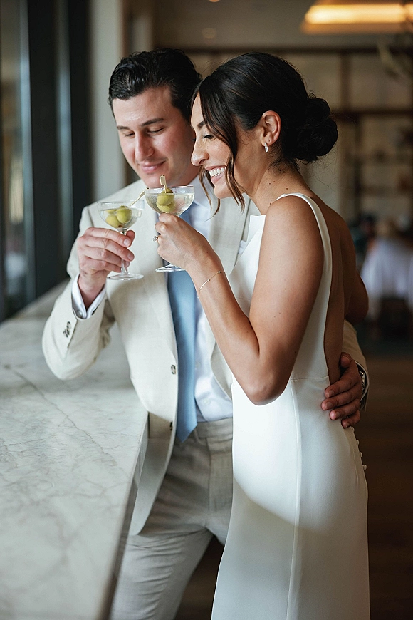 Wedding toast as bride and groom clink martini glasses with olive picks at a bar counter in soft window light during reception