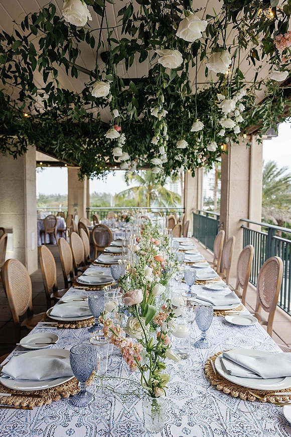 Reception tablescape with a long banquet table setup, blue goblets, patterned linens, taper candles, and hanging white roses under terrace lights