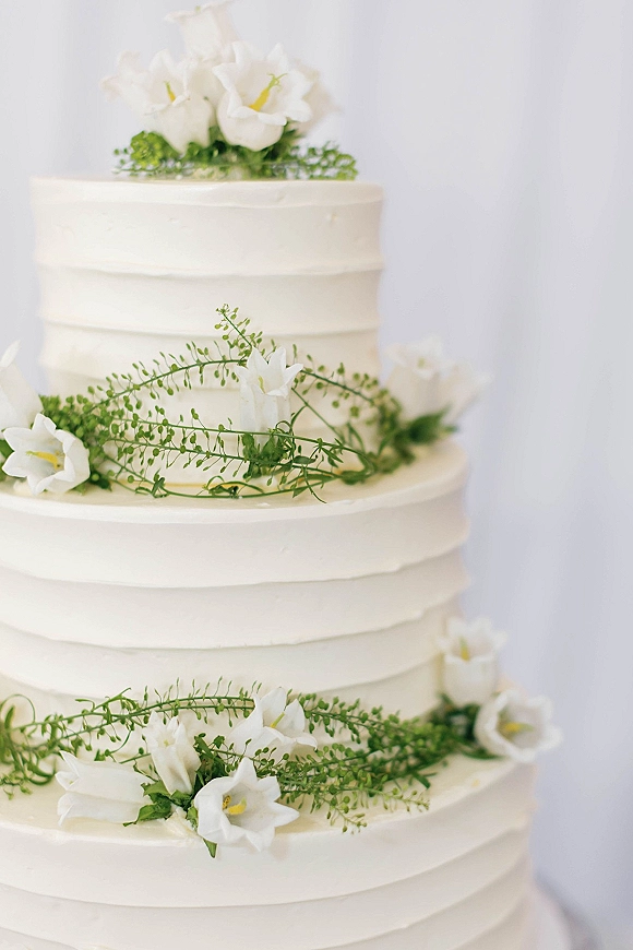Wedding cake with two tier wedding cake design, smooth white frosting and floral topper with white flowers and greenery against a neutral backdrop
