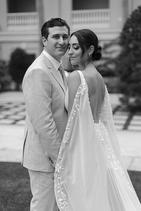 Couple portrait of bride and groom, bride looking over shoulder in backless dress with embroidered veil, in formal courtyard walkway