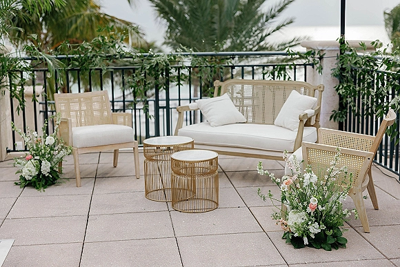 Wedding lounge setup with rattan outdoor sofa and white cushions, round coffee tables, and florals on a terrace overlooking ocean and palms