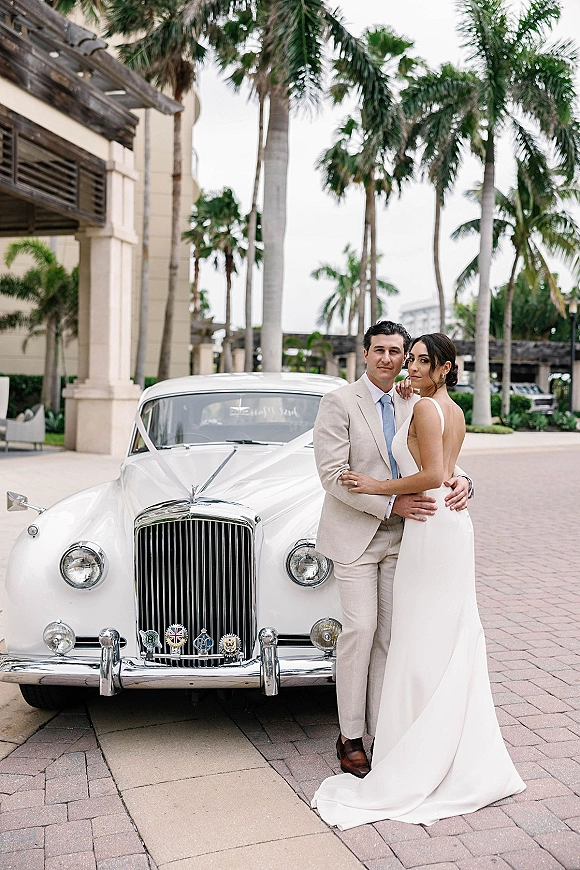 Couple portrait of bride and groom hugging beside a white vintage car, her satin dress train flowing on a palm-lined resort driveway