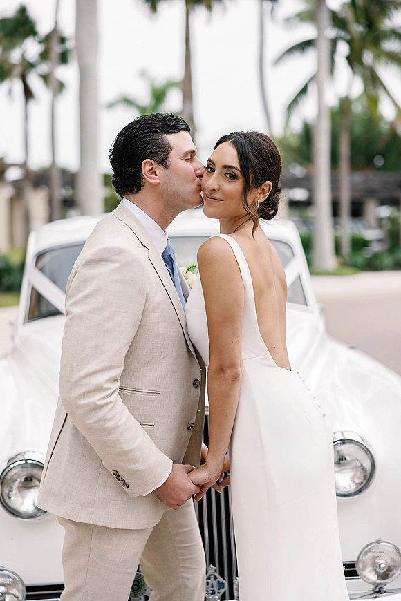 Couple portrait beside a vintage wedding car, groom kissing bride’s cheek as she shows an open-back gown on a palm-lined street