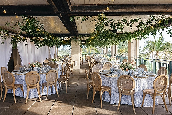 Reception tablescape with outdoor reception tables dressed in blue patterned linens, rattan chairs, floral centerpieces, and string lights on a covered patio