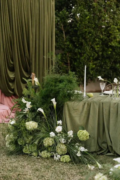 Reception tablescape with an olive green wedding tablescape, calla lilies and taper candle, set on a lawn beside a garden hedge backdrop