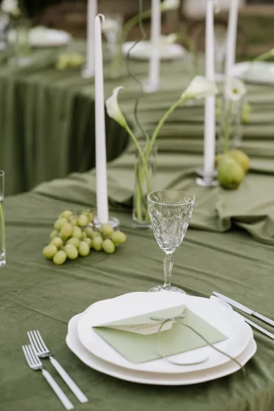 Reception tablescape with sage green tablescape linens, white taper candles, crystal goblets, calla lilies, and green fruit on a banquet table