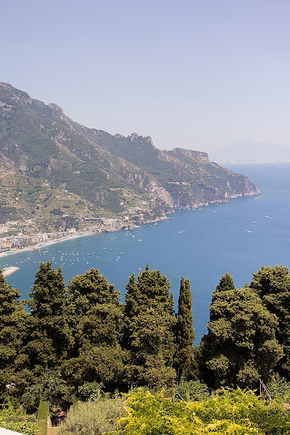 Coastal landscape with a mountain coastline view showing cliffside sea, a seaside town and boats below, framed by trees under clear sky