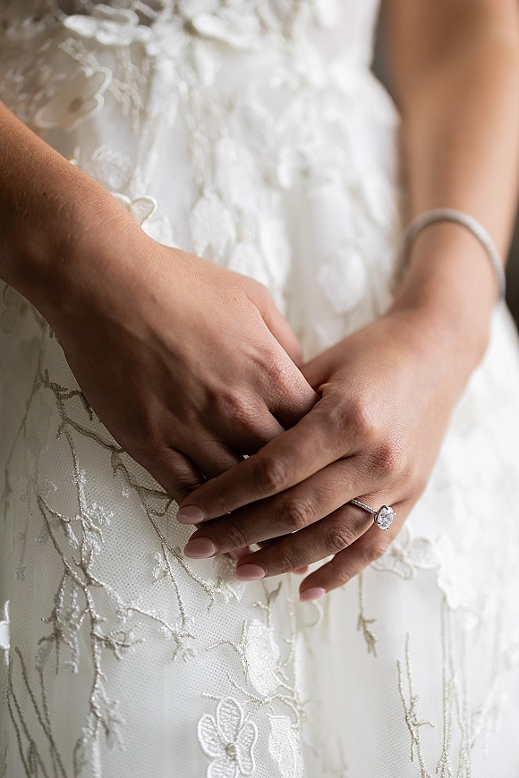 Engagement ring close-up of a solitaire engagement ring on manicured hand, diamond sparkling beside lace appliqué gown in soft indoor light