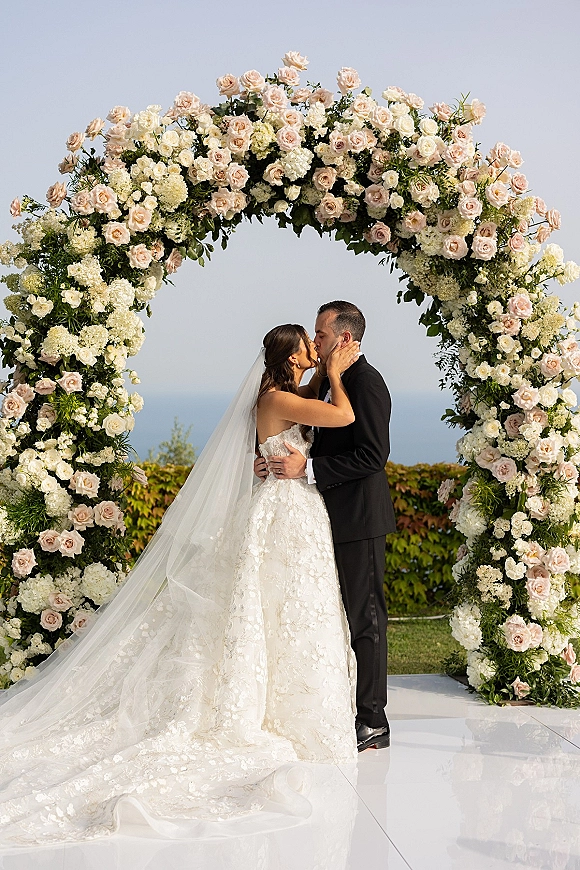 Wedding kiss portrait of bride and groom kissing under a rose and hydrangea floral arch, ocean view behind, veil and tuxedo flowing in breeze