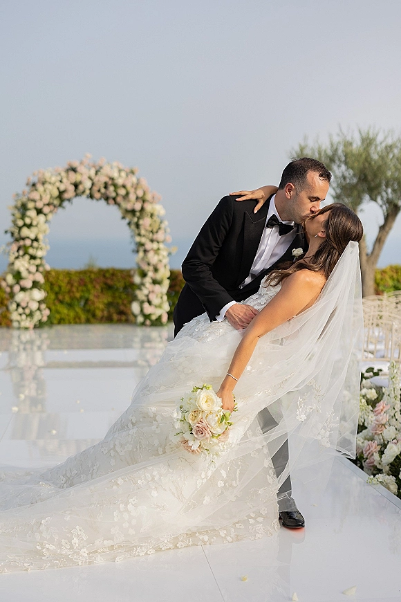 Wedding kiss portrait of groom dipping the bride beneath a floral arch, her veil flowing as greenery and a distant ocean frame the aisle