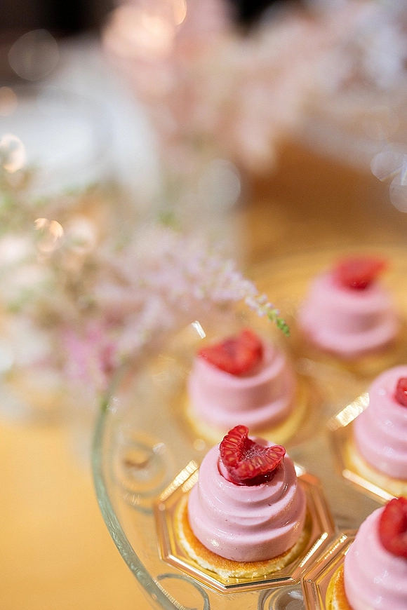 Wedding desserts arranged as mini cakes with pink frosting and raspberries on a glass platter atop a gold table with soft bokeh lights
