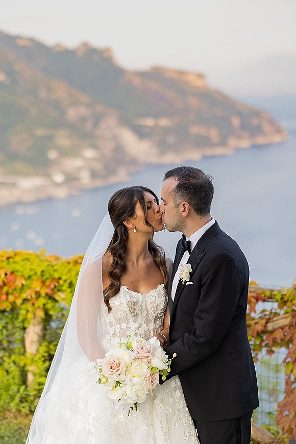Wedding kiss portrait of bride and groom kissing, bride in lace gown with veil and bouquet on coastal cliffs above ocean and hills