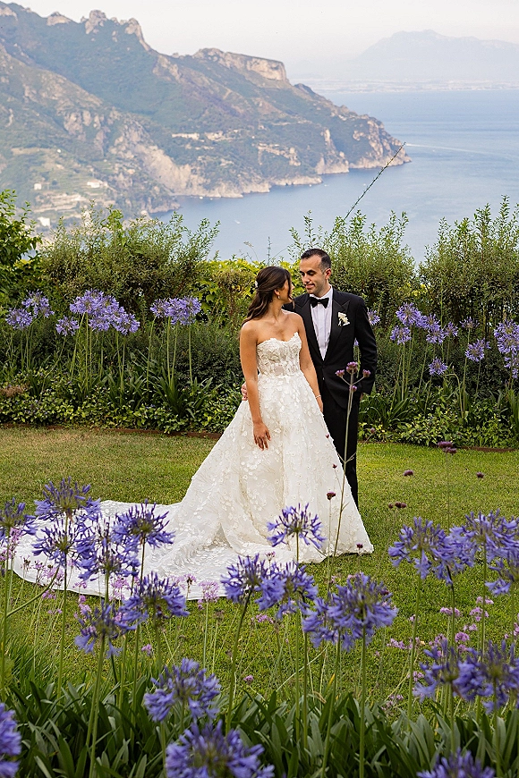 Couple portrait of bride in strapless lace dress with long train and groom in tuxedo, embracing on lawn by ocean and mountains backdrop