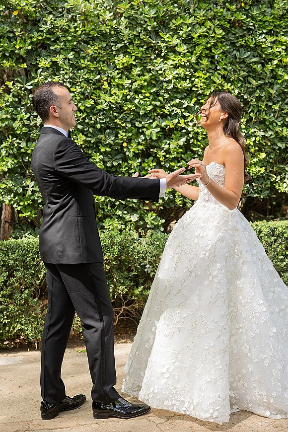 First look moment as bride in a strapless lace wedding dress meets groom in tuxedo on a stone garden path by green hedges