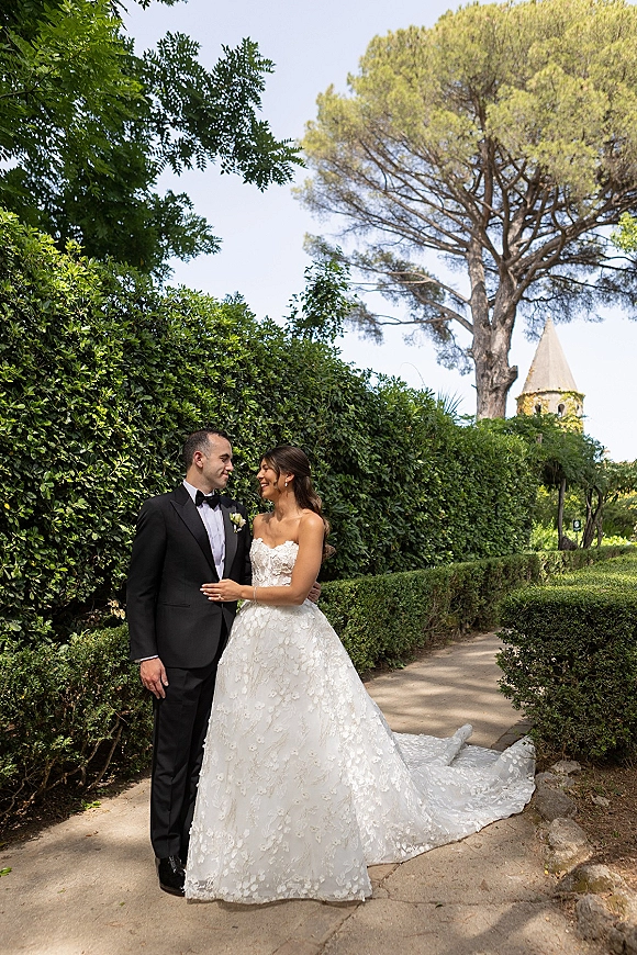 Couple portrait of bride in a strapless lace gown with long train and groom in tuxedo on a garden path with hedges and stone tower