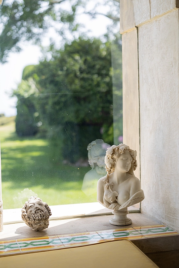 Wedding venue details on a tiled windowsill with a classical bust statue and stone head sculpture by a large window overlooking garden lawn