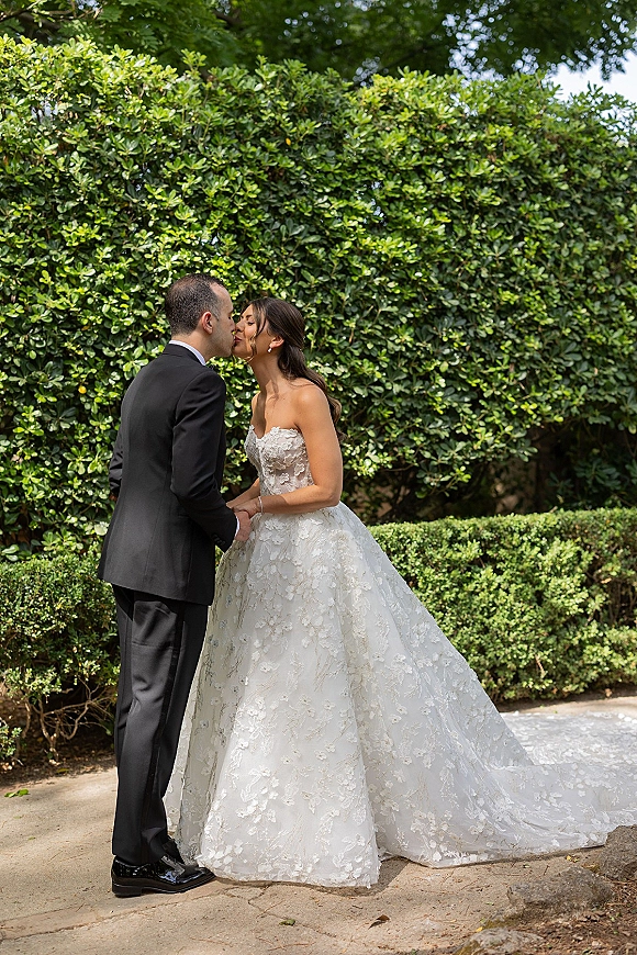 Wedding kiss portrait of bride and groom kissing, holding hands on a stone garden path, her strapless lace dress with long train