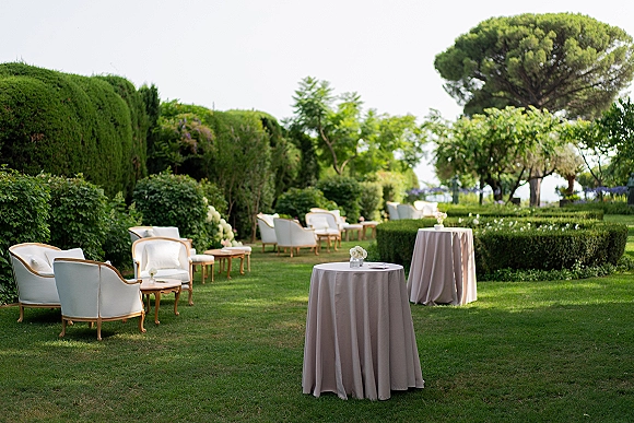 Outdoor lounge setup with wedding cocktail hour lounge chairs, linen-draped cocktail tables, and small florals on a manicured garden lawn