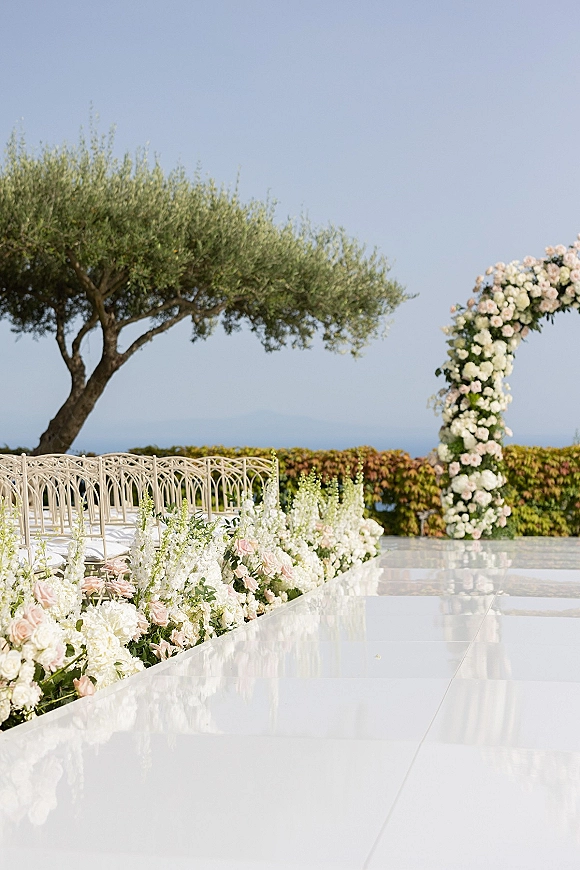 Ceremony aisle design with an outdoor wedding aisle, white runner and blush-white rose florals leading to a white arch with ocean view