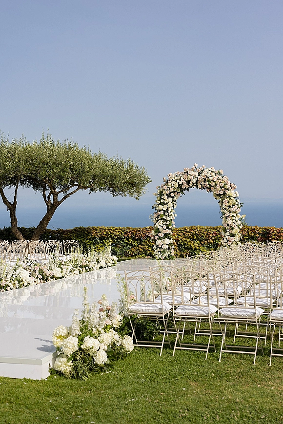 Ceremony setup with a round floral wedding arch and flower-lined aisle, white chairs on a lawn with ocean view backdrop