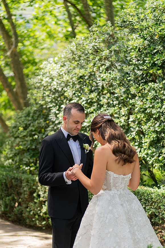 First look moment as bride and groom holding hands on a shaded garden path, showing her strapless lace dress and his black tuxedo
