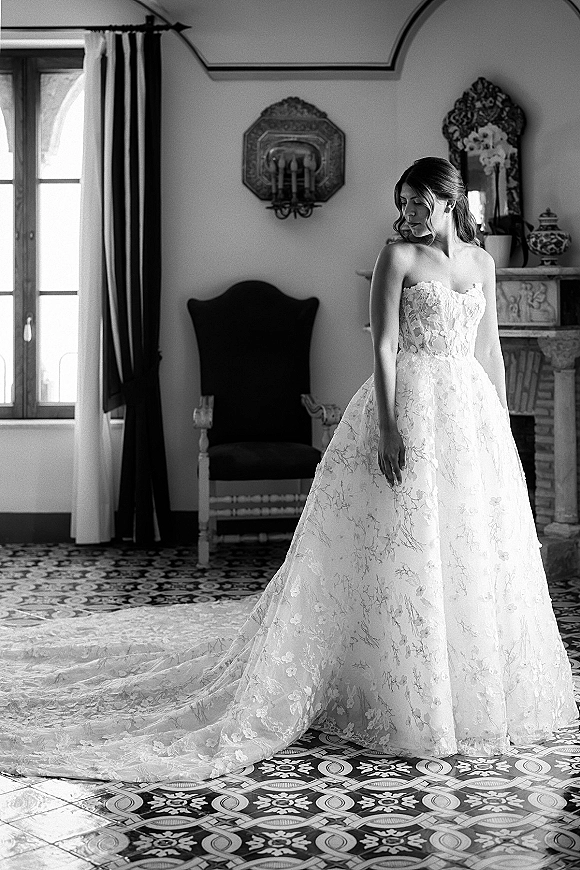 Bridal portrait of a bride in a strapless lace wedding dress with long train, half-up hair, standing by an arched window in a vintage room