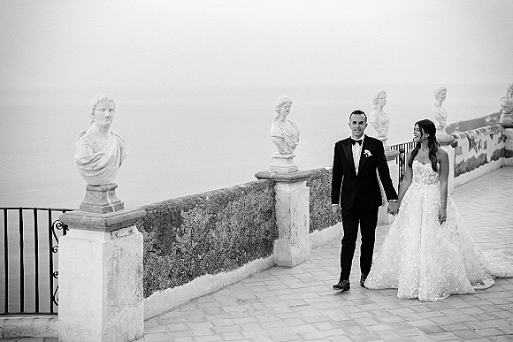 Couple portrait of bride and groom holding hands, her strapless lace dress train flowing on a stone terrace with ocean view