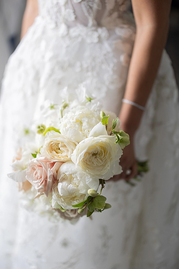 Bridal bouquet of white and blush roses with peonies and greenery held against a lace wedding dress in a soft indoor setting