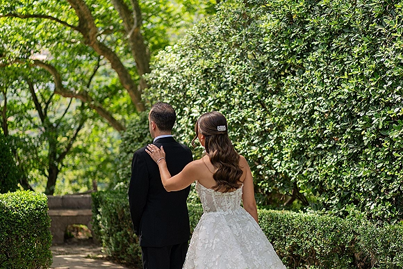Wedding first look as bride taps groom shoulder from behind, wearing strapless lace gown, on garden stone steps with hedges and trees