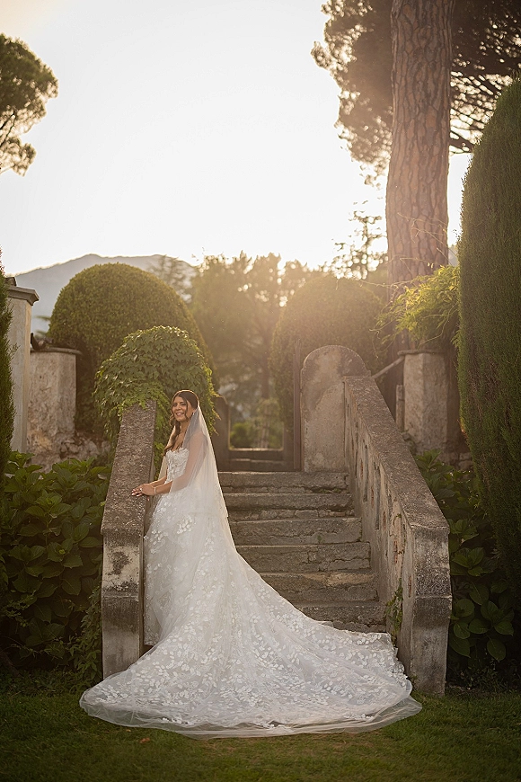 Bridal portrait of a bride in a strapless lace gown with cathedral veil and long train on a sunlit stone staircase by hedges