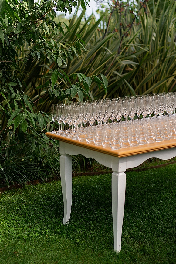 Champagne toast setup with champagne flute display on a cocktail table, arranged on a green garden lawn with shrubs behind