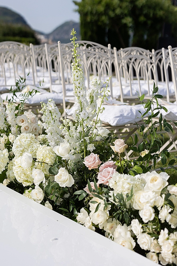 Ceremony aisle flowers in a lush aisle floral arrangement with white and blush roses beside white metal chairs in an outdoor garden setting