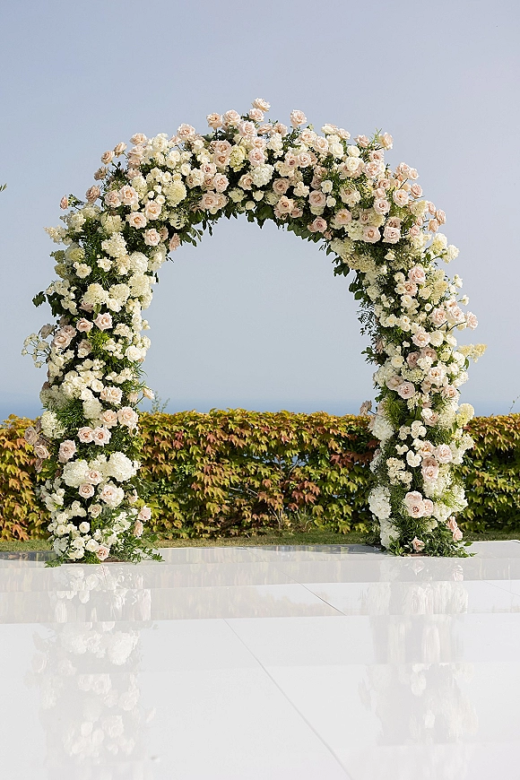 Wedding floral arch with round wedding arch flowers in blush and white roses and greenery, set on a glossy white aisle by the ocean horizon
