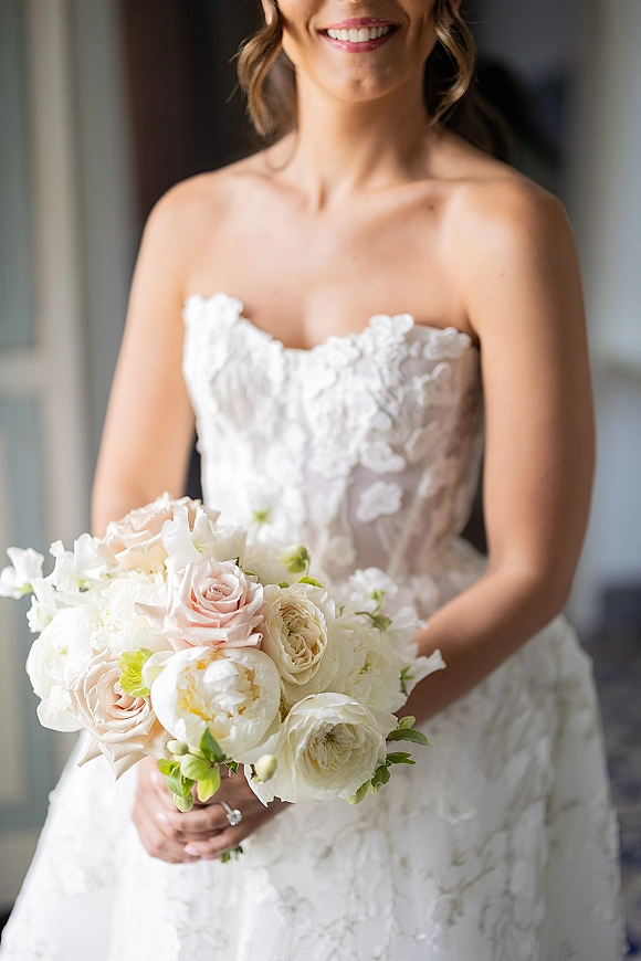 Bridal portrait of a bride holding bouquet in a strapless lace wedding dress, showing blush roses and white peonies in a doorway-lit room