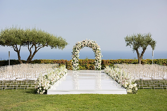 Ceremony setup with a round floral arch, blush and white aisle florals, and white chairs on a lawn overlooking the ocean backdrop