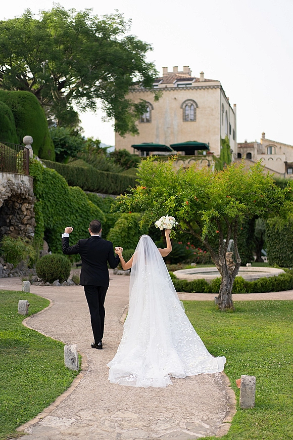 Newlywed couple walking away holding hands, bride raising bouquet with long veil and groom in black suit on a stone path by a villa fountain