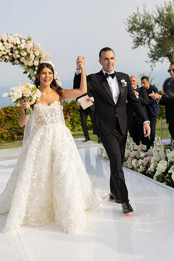 Wedding recessional as bride and groom walk the aisle hand in hand, she holds a white bouquet, guests applaud with ocean view behind