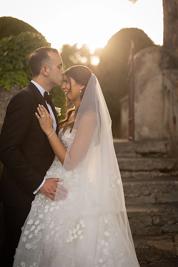 Wedding couple portrait of groom kissing bride’s forehead as they embrace on sunlit stone steps, her veil softly backlit by greenery
