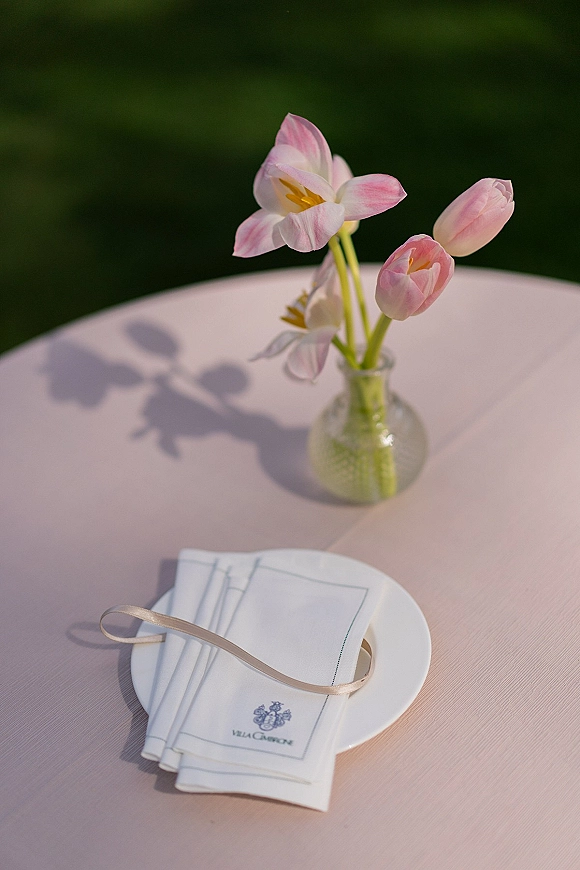 Reception table decor with bud vase centerpiece holding pink tulips on a blush tablecloth, folded napkin tied with ribbon, greenery behind