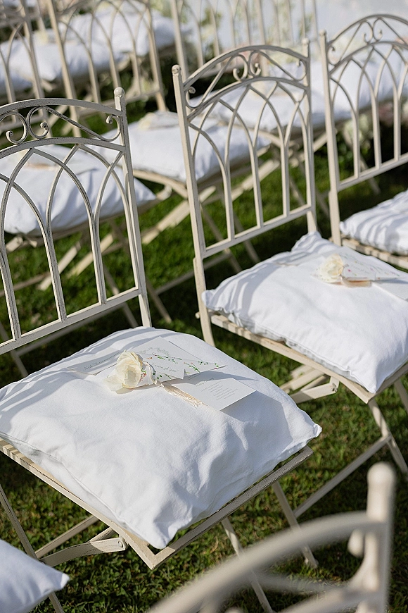 Ceremony seating with outdoor ceremony chairs on a grass lawn, each with white cushions, programs, and a white rose tied with ribbon